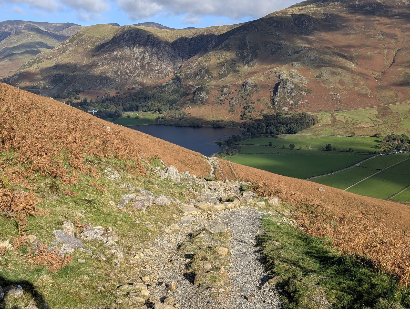 A stony path dropping through autumn bracken towards the head of Buttermere, green fields by the lake and craggy fellsides rising beyond