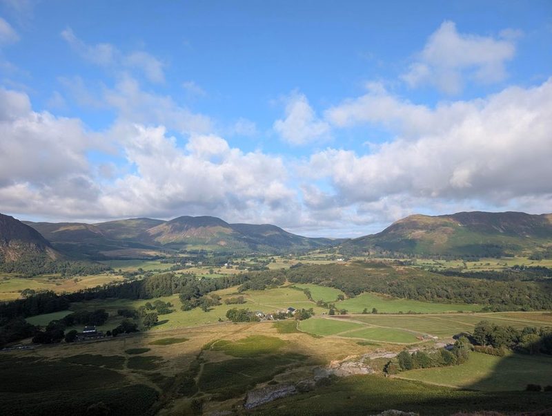 Looking back down over the green valley with its farms, walls and woods, and a line of fells catching the early sun along the far side