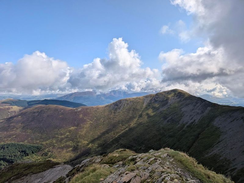 A steep grassy ridge falling away with a thin path along its crest, plantation and farmland in the valley beyond and cloud shadows crossing the fells