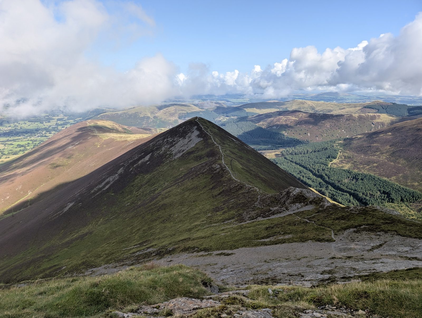 A steep grassy ridge falling away with a thin path along its crest, plantation and farmland in the valley beyond and cloud shadows crossing the fells