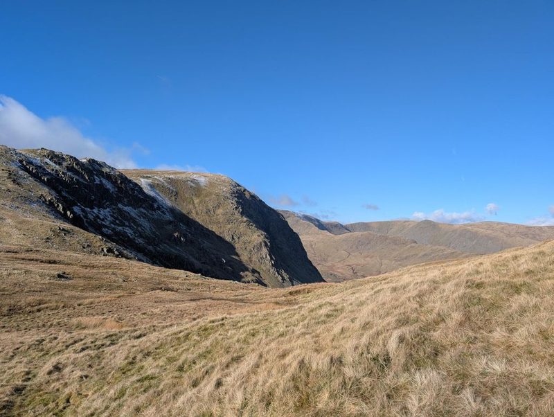 A dark crag streaked with snow above a wide sweep of bleached winter grass, with a deep valley and long rounded ridges beyond