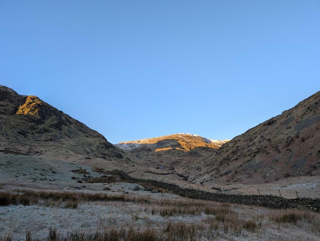 First sun catching the snow-dusted tops at the head of a frosted valley, with a drystone wall running up the pale winter grass