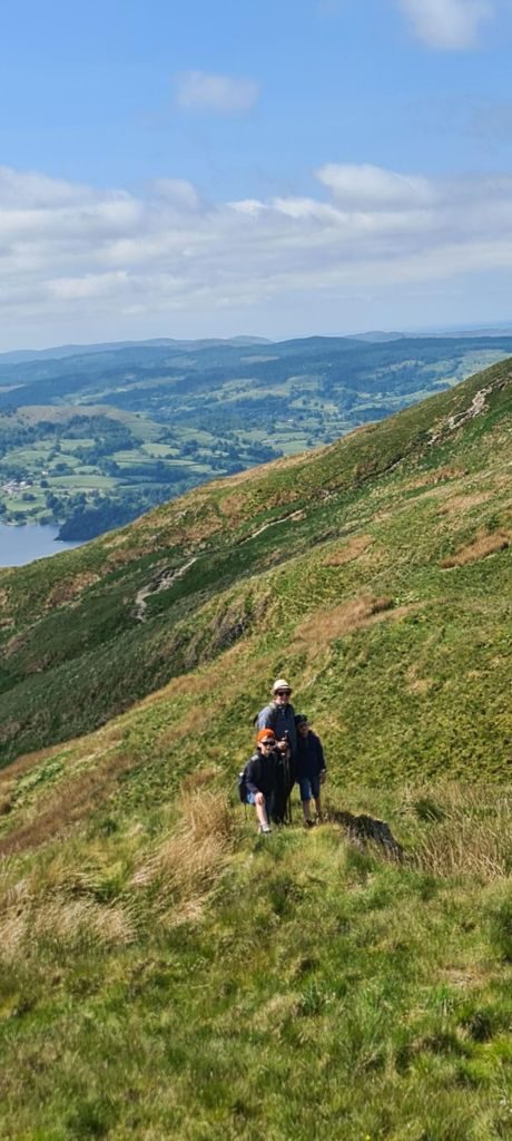 Three walkers coming up a narrow path across a steep grassy fellside, with the valley's fields and the head of the lake far below