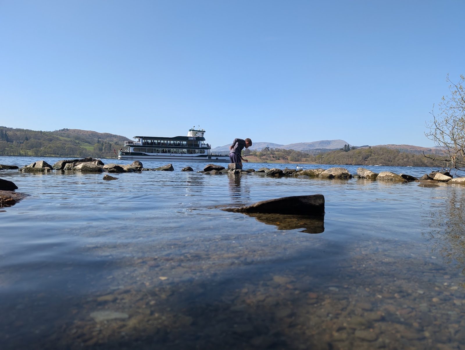 A child bends over stones at the water's edge of Windermere as a lake cruise boat passes, with wooded shores and fells beyond under a clear sky